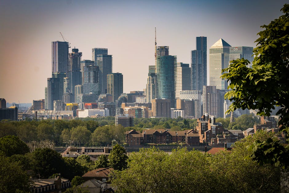 A wide view of a modern city skyline featuring high-rise office buildings and residential towers, with some structures under construction and cranes visible. In the foreground, there are green trees and residential houses with pitched roofs, reflecting a contrast between urban development and natural greenery. The scene captures a clear day with bright lighting, typical of a city located near a park or green space, suitable for home relocation or furniture transport services by Greenwich Man and Van, as referenced on the Greenwich webpage about avoiding hidden removal charges in Greenwich.