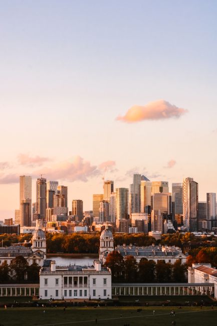 The image depicts a city skyline during sunset with tall modern office buildings and skyscrapers, some with glass facades reflecting the warm light. In the foreground, there is a historic white building with classical architecture, featuring multiple windows and a central dome, situated within a park or open green space that has neatly maintained lawns and a white fence running along its boundary. The scene is captured from an elevated vantage point, showing the spacious outdoor area in front of the historic building, which may be used for events or gatherings. The lighting creates a soft, warm glow across the cityscape, highlighting the contrast between the contemporary high-rises and the traditional architecture. This scene could be part of a house removal or interior moving context, with [COMPANY_NAME] potentially involved in logistics or transport services for urban relocations, emphasizing professional furniture transport and packing in a metropolitan setting.