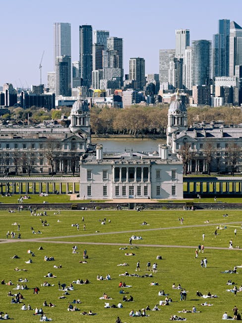 A historic white building with classical architectural features, including columns and a central entrance, is situated on a large grassy park area where numerous people are sitting, relaxing, and walking on the green lawns. In the background, a calm river separates the park from a modern cityscape composed of high-rise office buildings and skyscrapers with glass facades. The scene is captured during daylight hours under clear skies, and the area appears busy with individuals engaged in outdoor leisure activities. This setting exemplifies a popular urban park used for outdoor recreation and features elements relevant to house removals and home relocation services, with the modern buildings highlighting the importance of logistical planning for furniture transport and packing during moves, similar to the services offered by Greenwich Man and Van.