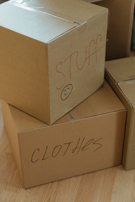 Close-up view of two cardboard boxes stacked inside a home, used for packing during a house removal. The top box has the word 'STUFF' written in red marker with a small smiley face underneath, while the lower box is labeled 'CLOTHES' in black marker. The boxes are sealed with packing tape and placed on a wooden floor, surrounded by other similarly packed boxes, indicating an organized packing process part of a furniture transport or home relocation. The scene reflects the careful packing and labeling carried out by Greenwich Man and Van during their removals and moving services, specifically for tight access situations as described in the 'Cutty Sark Greenwich removals packing tips for tight access' context.
