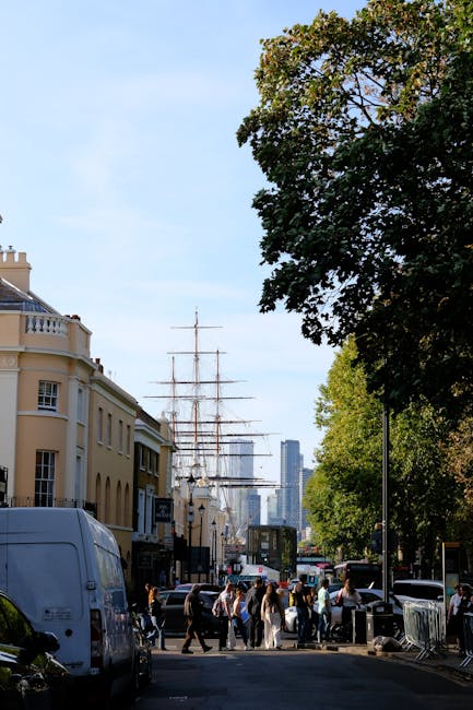 The image shows a busy street scene in Greenwich with a tall sailing ship moored nearby, partially visible through the trees on the right. On the left side, there are several multi-storey residential buildings with cream and light brown facades, featuring windows and balconies. A white van is parked on the street, while pedestrians are walking along the pavement, some in groups and others alone, indicating a lively urban environment. In the background, modern high-rise buildings can be seen against a clear blue sky. The scene is well-lit with natural daylight, suggesting a bright, sunny day. This setting reflects an active neighborhood that could be involved in home relocations or furniture transport, which aligns with the services offered by Greenwich Man and Van, especially in the context of packing tips for tight access during house removals.