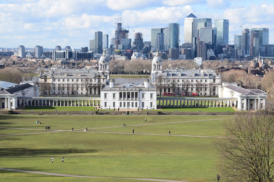 A wide view of a large, historic white building with classical architecture, including domed towers and colonnades, situated within a spacious green park area with walking paths and several people scattered across the grass. The building appears to be set against a backdrop of modern high-rise skyscrapers and office buildings under a partly cloudy sky. In the foreground, the park's open lawns are dotted with visitors, some walking and others sitting. The image suggests a peaceful setting with an emphasis on spaciousness, typical of a location like Greenwich Park. Greenwich Man and Van offers house removals and relocation services, which often involve navigating access challenges similar to those shown by this large historic building within a busy urban environment, highlighting the importance of professional furniture transport and careful packing during home relocation projects.