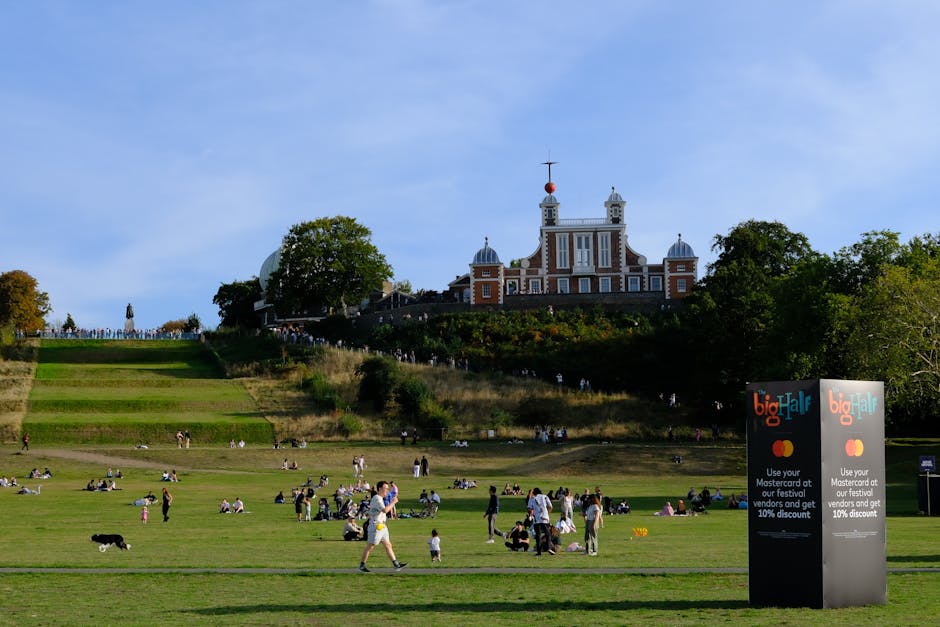 A wide view of Greenwich Park with a large, ornate building at the top of a hill under a clear blue sky. The park's sloping grassy landscape is populated with groups of people sitting, walking, and children playing. On the right side of the image, there is a black sign with the text 'big half' and promotional information near the park entrance. The foreground features a paved pathway with several individuals and a dog in motion, while the background shows trees lining the hillside. The scene captures a lively outdoor environment, illustrating park visitors and the accessible terrain, relevant to home relocation or moving services provided by Greenwich Man and Van, particularly those facing access challenges during house removals.