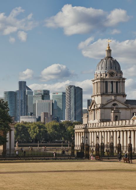 A view of a historic building with a domed tower and classical architectural details, situated in a park with a paved walkway and benches in the foreground. Behind the building, there is a modern city skyline featuring tall glass skyscrapers and office buildings. The scene is under a partly cloudy sky with some patches of blue, and the park area has trees and open grass. This image relates to house removals and moving logistics, illustrating the contrast between historic and modern architecture, which may be relevant to relocation services offered by Greenwich Man and Van in SE10, focusing on home relocation and furniture transport.