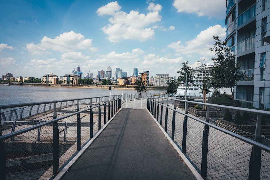 View of a metal pedestrian walkway with protective fencing on both sides, extending over a river in an urban area. The pathway is situated next to modern residential buildings with glass balconies and large windows on the right side. In the background, a city skyline featuring various high-rise buildings and cranes is visible under a partly cloudy sky. The scene is well-lit by natural daylight, emphasizing the outdoor environment typical of house removals or parts of a moving process near riverside properties in Greenwich. Greenwich Man and Van's removals service often includes transporting and loading possessions across such locations, supported by logistics that involve access routes like this walkway.