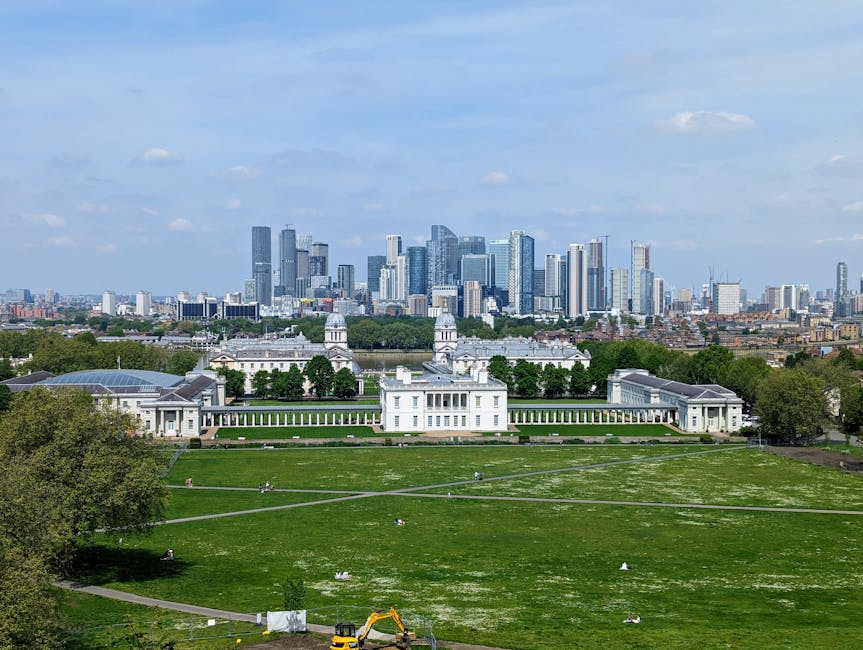 A view of a historic building with a domed tower and classical architectural details, situated in a park with a paved walkway and benches in the foreground. Behind the building, there is a modern city skyline featuring tall glass skyscrapers and office buildings. The scene is under a partly cloudy sky with some patches of blue, and the park area has trees and open grass. This image relates to house removals and moving logistics, illustrating the contrast between historic and modern architecture, which may be relevant to relocation services offered by Greenwich Man and Van in SE10, focusing on home relocation and furniture transport.