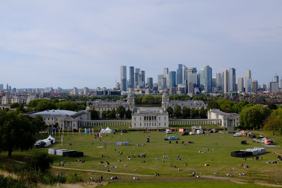 A wide-angle view of a large outdoor park with a historic white building located in the middle ground, surrounded by green lawns and scattered visitors sitting or walking across the field. In the foreground, there are various cardboard boxes, wooden furniture, and moving blankets indicating furniture and packing materials used during a home relocation process. To the left, a black trolley and a stack of plastic wrapping materials are visible, while in the background, a delivery van is parked near the edge of the property. Beyond the park, a modern city skyline with tall glass skyscrapers dominates the horizon under a partly cloudy sky, suggesting an urban setting where house removals by Greenwich Man and Van may occur. The environment is well-lit with natural daylight, emphasizing the scene's clarity and activity related to packing, furniture transport, and moving logistics, illustrating typical elements encountered in professional removals service activities.