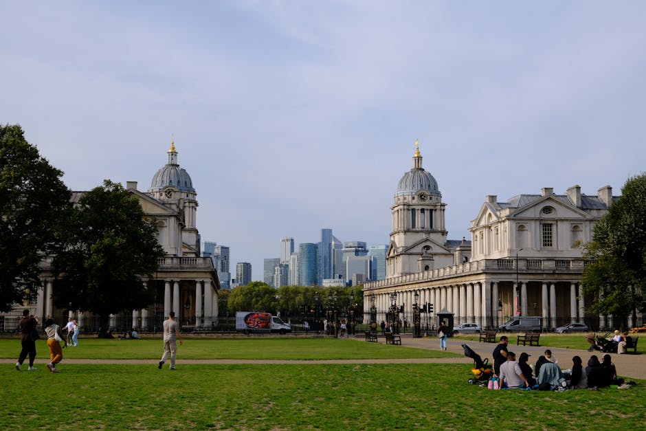 A wide view of a park in Greenwich featuring two historic white stone buildings with classical architectural elements, including columns and domed roofs topped with golden finials. The buildings are situated on the edge of the park with the city skyline visible in the background, consisting of modern high-rise office buildings and skyscrapers. The park has a well-maintained grassy area, with several people sitting on blankets and on the grass, some walking or cycling along pathways. A few trees are positioned around the park, providing shade, and a van with promotional graphics is parked along the street behind the buildings. The scene is illuminated by natural daylight with partly cloudy skies overhead, capturing a calm and organized outdoor space that could be part of a home relocation process involving moving through Greenwich, as handled by Greenwich Man and Van.