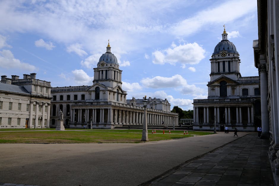 A wide view of a park in Greenwich featuring two historic white stone buildings with classical architectural elements, including columns and domed roofs topped with golden finials. The buildings are situated on the edge of the park with the city skyline visible in the background, consisting of modern high-rise office buildings and skyscrapers. The park has a well-maintained grassy area, with several people sitting on blankets and on the grass, some walking or cycling along pathways. A few trees are positioned around the park, providing shade, and a van with promotional graphics is parked along the street behind the buildings. The scene is illuminated by natural daylight with partly cloudy skies overhead, capturing a calm and organized outdoor space that could be part of a home relocation process involving moving through Greenwich, as handled by Greenwich Man and Van.
